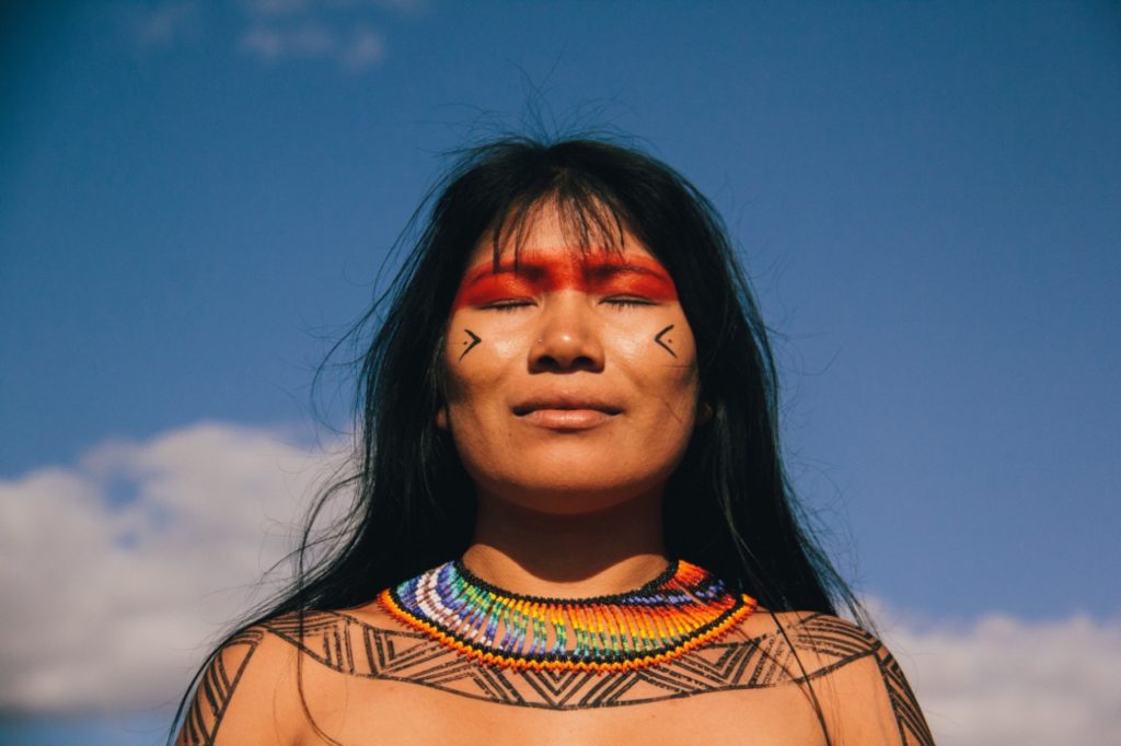 Mulher jovem indígena com pinturas corporais no rosto, colo, ombros e braços. Ela tem cabelos longos, escuros e sorri discretamente de olhos fechados. Em seu pescoço, colar colorido, feito de miçangas. Ao fundo, céu azul e nuvens brancas