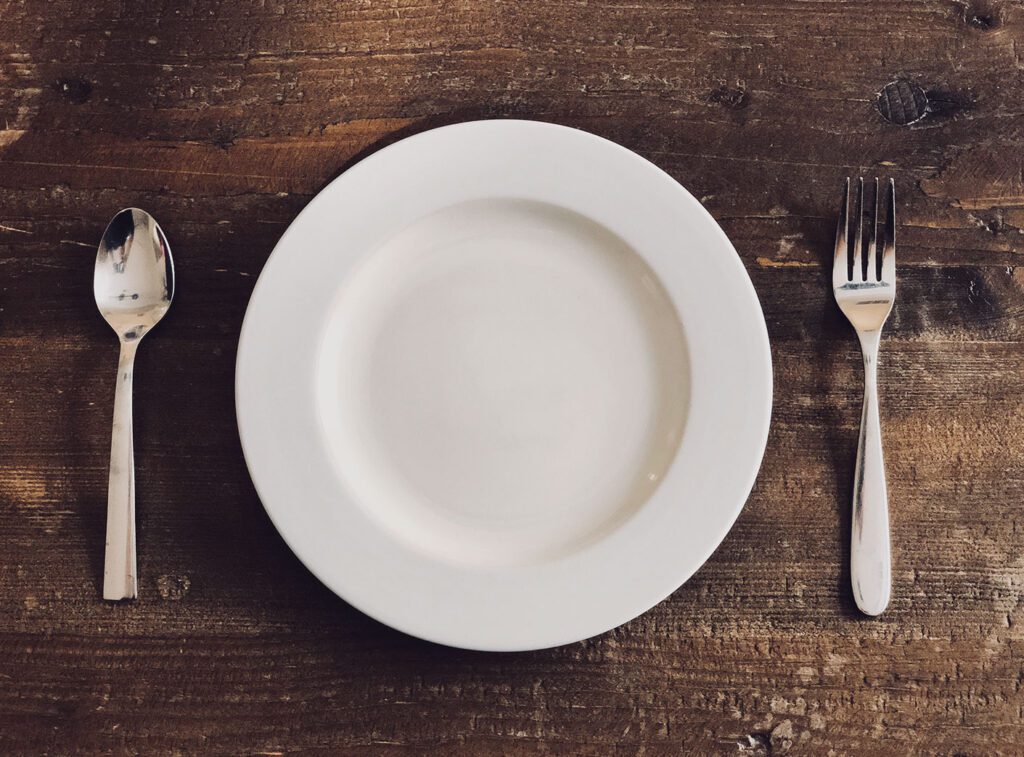 Prato branco, redondo e vazio sobre mesa marrom. À sua direita, garfo, prateado e liso. À esquerda, colher de igual aparência