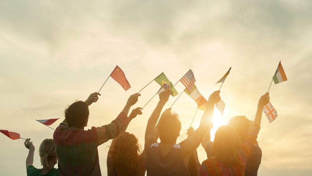 De costas, seis pessoas, de braços erguidos, seguram bandeiras de diferentes países e olham para o alto, mesma direção do reflexo dos raios de sol